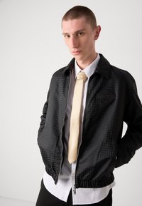 Veste zippée noire avec un motif texturé, finition matelassée et un détail de badge. Portée sur une chemise blanche et une cravate crème.