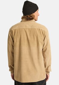 Beige kordfløjlsskjorte med en afslappet pasform, der har en rygsøm, knapmanchetter og en struktureret finish. Båret med en sort hue.