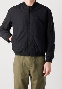 Veste bomber noire en tissu léger et texturé, avec col et poignets côtelés, fermeture éclair à l'avant et poches latérales. Portée avec un pantalon vert olive.