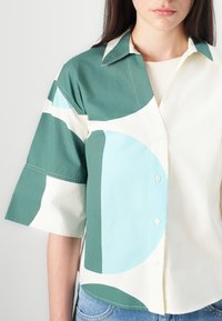 Chemise à manches courtes avec col, à motifs verts et bleu clair sur un tissu blanc, dotée d'un devant à boutons et d'un design décontracté.