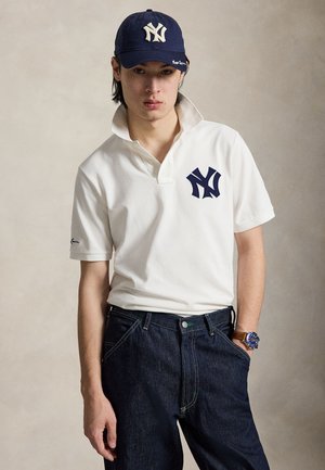 Fiatal férfi, fehér New York Yankees galléros pólót, tengerészkék sapkát, sötét farmert és karórát visel, egyszínű háttér előtt áll.