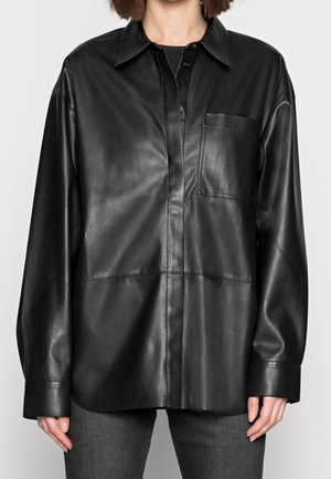 Kobieta nosząca luźną czarną skórzaną koszulę z przednią kieszenią i zapięciem na guziki, w połączeniu z ciemnoszarymi dżinsami.