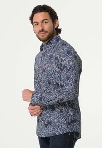 Blauw bloemenoverhemd met witte omlijningen, knoopsluiting, contrasterende rode knopen en lange mouwen. De stof lijkt glad en lichtgewicht te zijn.