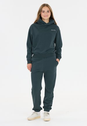 Jonge vrouw staand, gekleed in een donkerblauwe hoodie en bijpassende joggingbroek met witte sneakers, poseert met één hand in de zak tegen een witte achtergrond.