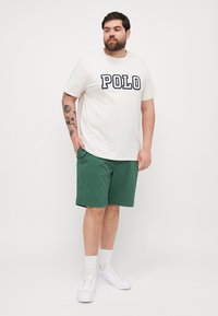 Tricou alb cu "POLO" în litere bleumarin, asociat cu pantaloni scurți verzi. De asemenea, se văd adidași albi și șosete texturate albe.