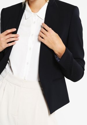 Nahaufnahme einer Person, die ein weißes Hemd mit Knöpfen, beige Hose und einen schwarzen Blazer trägt und das Revers des Blazers mit beiden Händen richtet.