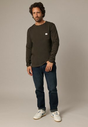 Olijfgroene blouse met lange mouwen en een borstzak, gecombineerd met donkerblauwe jeans en witte sneakers. De stof lijkt zacht en casual te zijn.