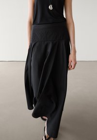 Sort maxi nederdel med en tekstureret talje, der har et blødt design og draperet stof for en afslappet silhuet. Parrret med en ærmeløs top.
