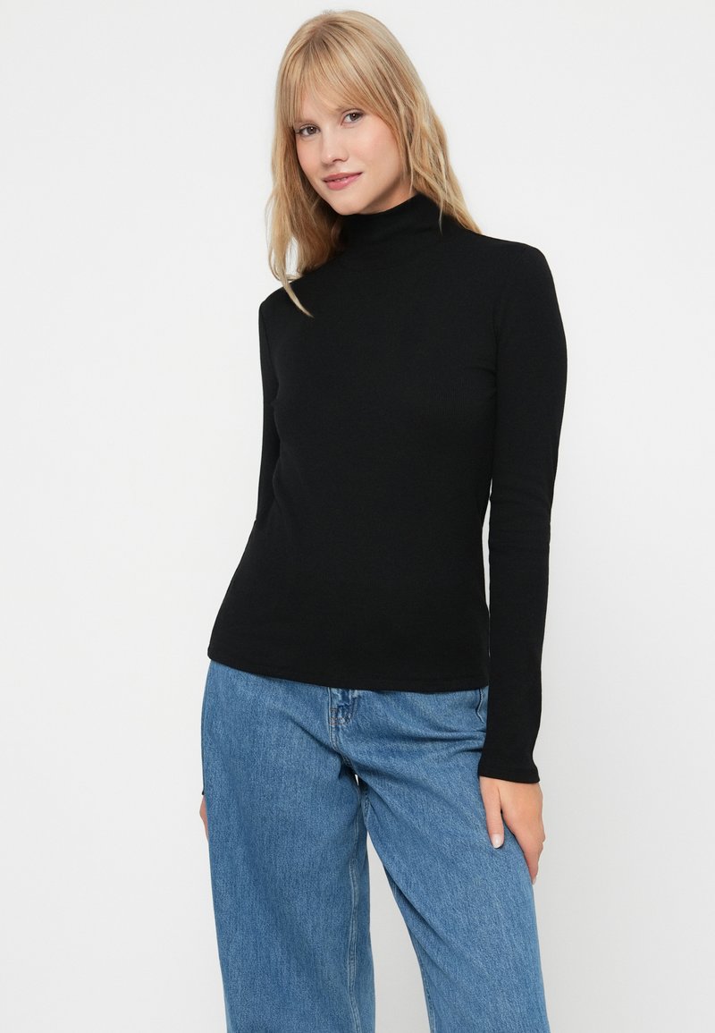 Maglione a collo alto nero, a maniche lunghe, con una trama a coste e un design aderente, abbinato a jeans in denim blu larghi. Sfondo semplice.