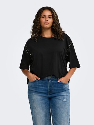 Femme aux cheveux bouclés portant un t-shirt noir à manches courtes avec de petits clous argentés et un jean bleu, les mains dans les poches.