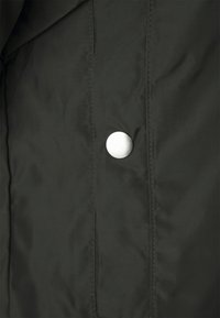 Svart, texturerad tyg med en glänsande silverfärgad tryckknapp. Plagget har noggrant sydda sömmar längs kanterna, vilket framhäver dess strukturerade design.