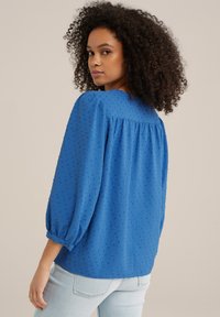 Blusa blu in tessuto testurizzato con maniche a sbuffo e polsini con bottoni. Il retro presenta un design arricciato e dettagli a motivo discreti.