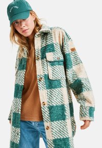 Gevlekte overhemdjasje in teal en crème, met een getextureerde, fuzzy stof, twee voorzakken en bruine knopen. Draagt over een bruine shirt.