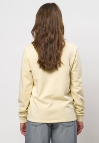 Persoon met lang golvend bruin haar, dragend een lichtgele trui en grijze spijkerbroek, met de rug naar de camera gericht op een effen witte achtergrond.