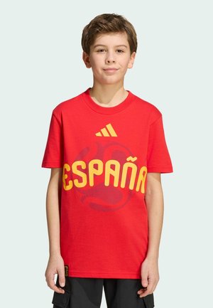 Chłopiec w czerwonej koszulce z żółtym napisem "España" i logo Adidas, stojący na jednolitym jasnym tle.
