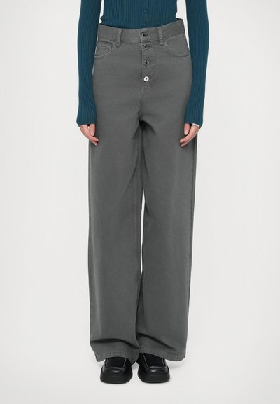 Persoană îmbrăcată cu pantaloni gri cu talie înaltă și picioare largi, cu închidere cu nasturi, top teal cu dungi, cu mânecă lungă și pantofi negri, stând în fața unui fundal alb.