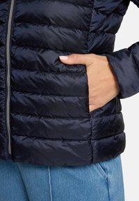 Doudoune bleu marine avec texture matelassée et fermeture éclair à l'avant. Comprend une poche latérale en tissu lisse et une coupe décontractée.