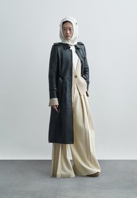 Μαύρο δερμάτινο παλτό με κουμπιά, περασμένο πάνω από ένα ανοιχτό πλεκτό τοπ και μια μπεζ γιλέκο, συνδυασμένο με φαρδιές μπεζ παντελόνες και ένα λευκό πλεκτό καπέλο.