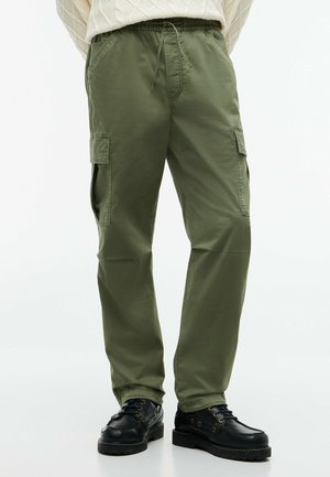 Mann trägt olivgrüne Cargohosen mit Seitentaschen, einen weißen Strickpullover und schwarze Leder-Schnürschuhe.