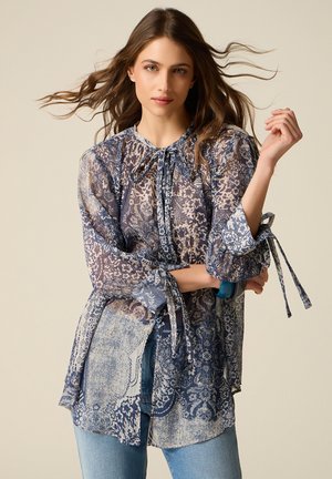 Frau mit langen braunen Haaren trägt eine transparente, gemusterte blaue Bluse mit gebundenen Manschetten und hellblaue Jeans, posiert vor einem neutralen Hintergrund.