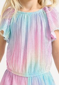 Giovane vestito con un abito pastello arcobaleno, con strisce verticali glitterate e maniche corte a balza, capelli che cadono sulle spalle.