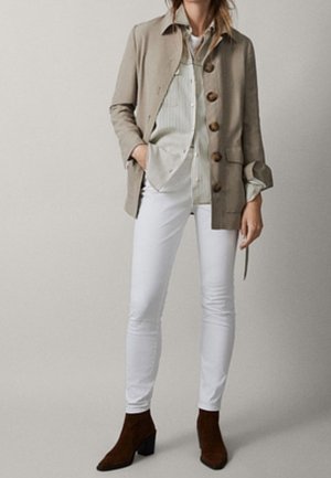 Frau trägt beige geknöpfte Jacke, hell gestreiftes Hemd, weiße enge Hose und braune Stiefeletten, steht vor einem schlichten Hintergrund.