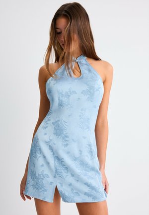 Femme portant une robe bustier sans manches bleu clair à fleurs avec une fente à l'avant, regardant vers le bas, les cheveux couvrant partiellement son visage.