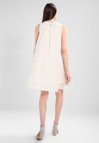 Frau mit langen dunklen Haaren, die ein ärmelloses weißes Lagenkleid und graue High Heels trägt, steht mit dem Rücken zur Kamera.