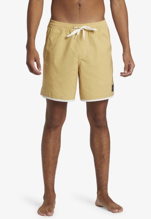 Gelbe Badeshorts mit einer weißen Kordel und Saum, versehen mit einem Patch-Logo auf der linken Seite, hergestellt aus leichtem, wasserabweisendem Material.