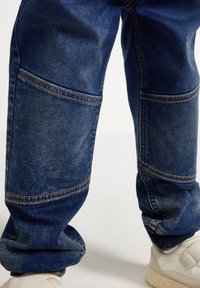 Denim joggers in dark blue with visible stitching, relaxed fit, and tapered cuffs, paired with light-colored sneakers.