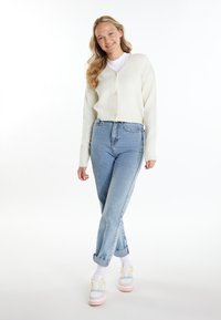 Cremfarbener, gestrickter Cardigan mit V-Ausschnitt und Knöpfen, kombiniert mit hellblauen, hochgeschnittenen Jeans und mehrfarbigen Turnschuhen.