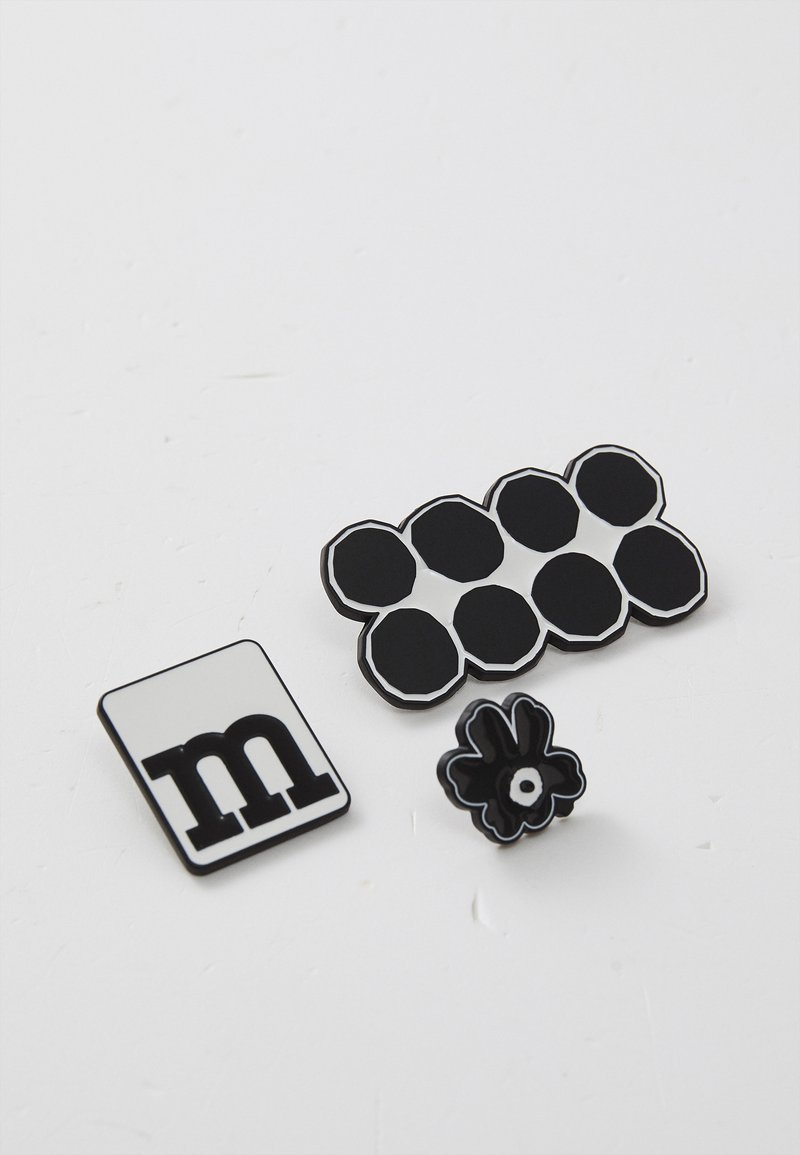 Three enamel pins: a rectangular pin with a black "m" on a white background, a hexagonal design with black and white, and a black flower pin with a white center.