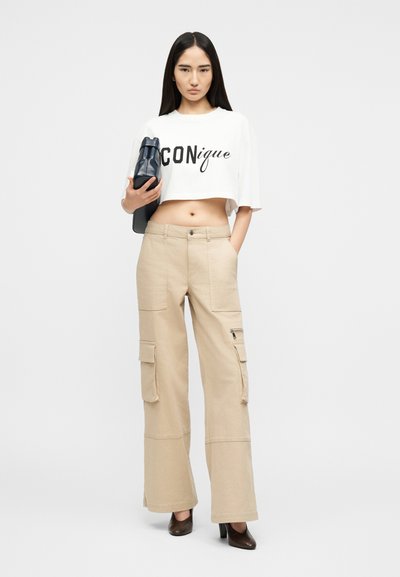 Νεαρή γυναίκα στέκεται φορώντας λευκό τοπ με το κείμενο «CONique», μπεζ cargo παντελόνια, μαύρα τακούνια, κρατώντας μια μαύρη τσάντα σε λευκό φόντο.