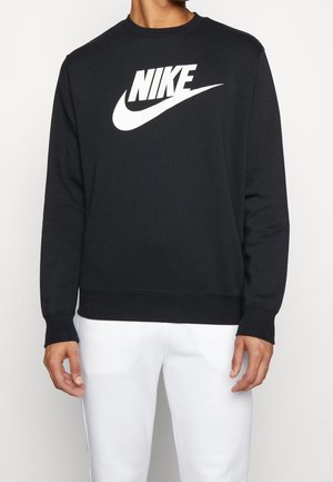 Mann trägt schwarzen Nike-Sweatshirt mit weißem Logo und weißen Hosen, steht vor einem einfarbigen hellen Hintergrund.