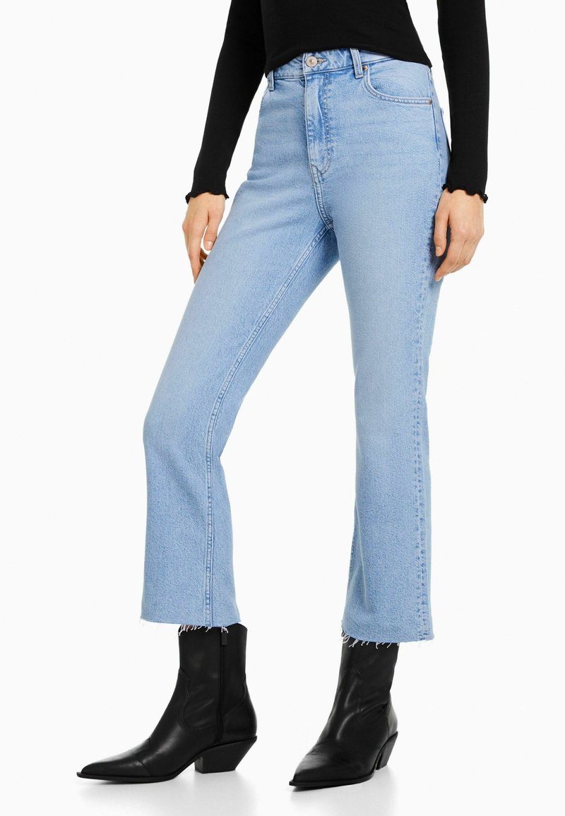 Femme portant un jean flare court bleu clair avec ourlets bruts, un haut noir à manches longues et des bottines noires à bout pointu.
