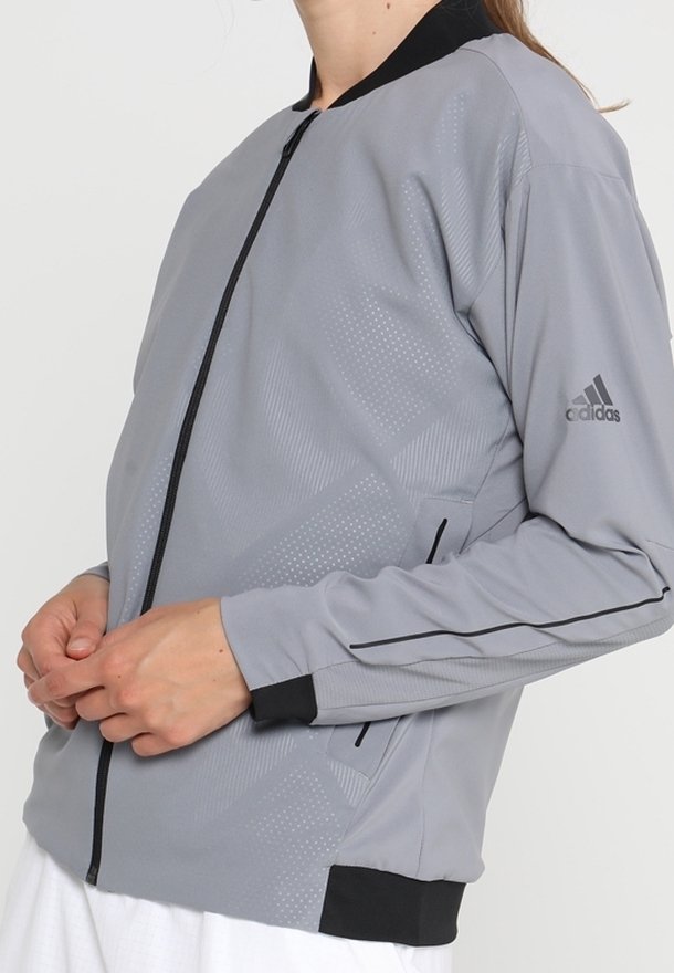 Grå sportjacka med en slät yta, svarta detaljer, dragkedjestängning och subtilt mönster. Har Adidas-logotyp på ärmen.