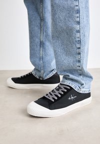 Mustat kangaskengät valkoisilla kumipohjilla, harmaat nauhat ja logotiedot. Yhdistetty vaaleansinisiin farkkuihin.