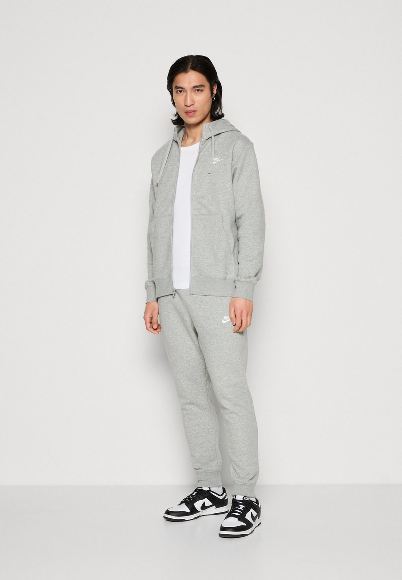 Ensemble sweat à capuche gris et jogger en tissu doux, avec fermeture éclair avant, poches et accents de logo. Associé à des baskets noires et blanches.
