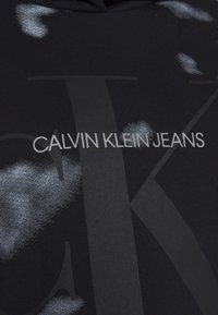Felpa nera con un grande logo grigio che recita "CALVIN KLEIN JEANS" e motivi di schizzo bianchi astratti sul tessuto.