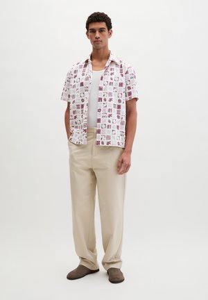 Mann steht in beigen Hosen, braunen Schuhen, weißem Tanktop und offenem Hemd mit kurzen Ärmeln und roten sowie weißen geometrischen Mustern.