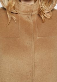 Helle braune Wolljacke mit Kapuze, Knopfverschluss und quadratischen Taschen, mit weicher Textur und klaren Linien.