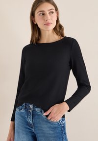 Top nero a maniche lunghe con coste e scollo rotondo. La texture del tessuto è liscia e leggermente elastica. Indossato con jeans di denim blu.