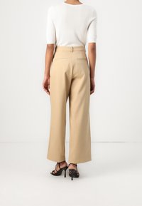 Beiga vida byxor med hög midja och bakfickor, i kombination med svarta remsandaler, sedd bakifrån mot en vit bakgrund.