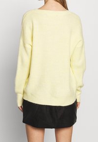Gelber gerippter Strickpullover mit überschnittenen Schultern, lockerer Passform und abgerundetem Saum, kombiniert mit einem schwarzen Rock. Weiche Textur ist offensichtlich.