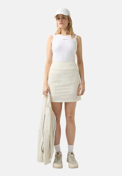 Witte geribbelde tanktop, lichtgekleurde A-lijn rok met een gestructureerd patroon, houdt een bijpassende jas vast, draagt een witte pet en beige sneakers.