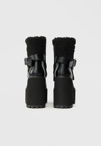 Melnas platformas potītes ar masīviem papēžiem, ādas siksnām ar sprādzēm un vilnas oderi augšējās apakšdaļās, parādītas no aizmugures.