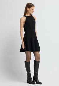 Zwarte mouwloze jurk met een uitlopende rok, gemaakt van gebreide stof. Gecombineerd met hoge zwarte leren laarzen met een gouden accent.