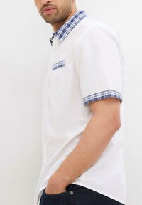 Camicia bianca a maniche corte con colletto e tasca a fantasia tartan blu. Tessuto morbido con una vestibilità comoda, abbinata a jeans scuri.