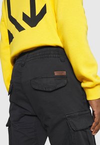 Zwarte cargobroek met een gestructureerde afwerking, voorzien van een elastische tailleband en zijzakken, gecombineerd met een felgele sweatshirt.