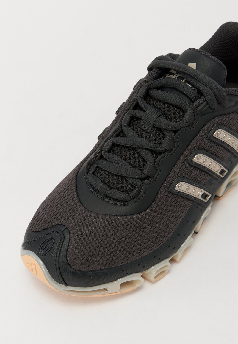 Zapatilla deportiva negra con parte superior de malla, cierre de cordones, tres franjas metálicas y una suela de goma acolchada con cámaras de aire visibles.